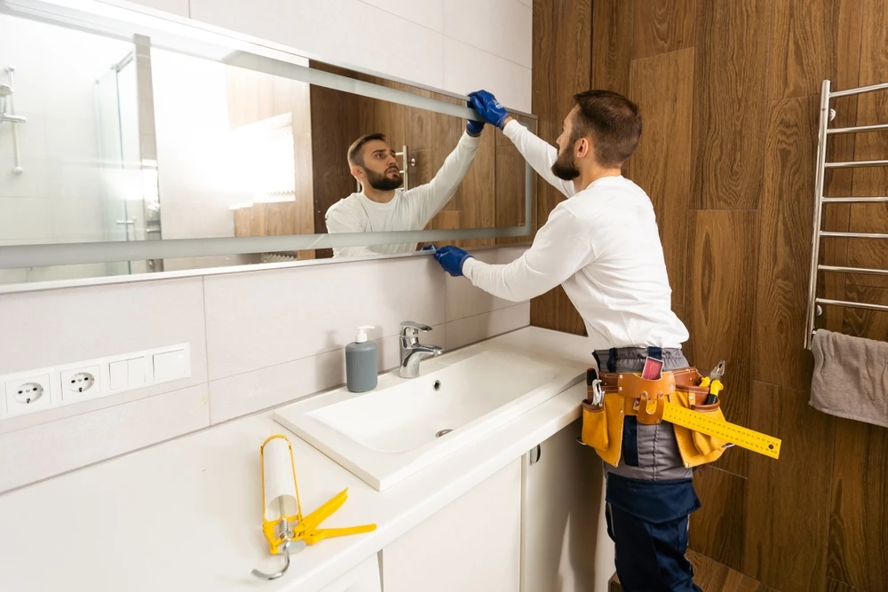 A handyman wearing blue gloves and a tool belt installs or adjusts a large rectangular mirror above a white bathroom vanity. A caulking gun sits on the counter in the foreground.