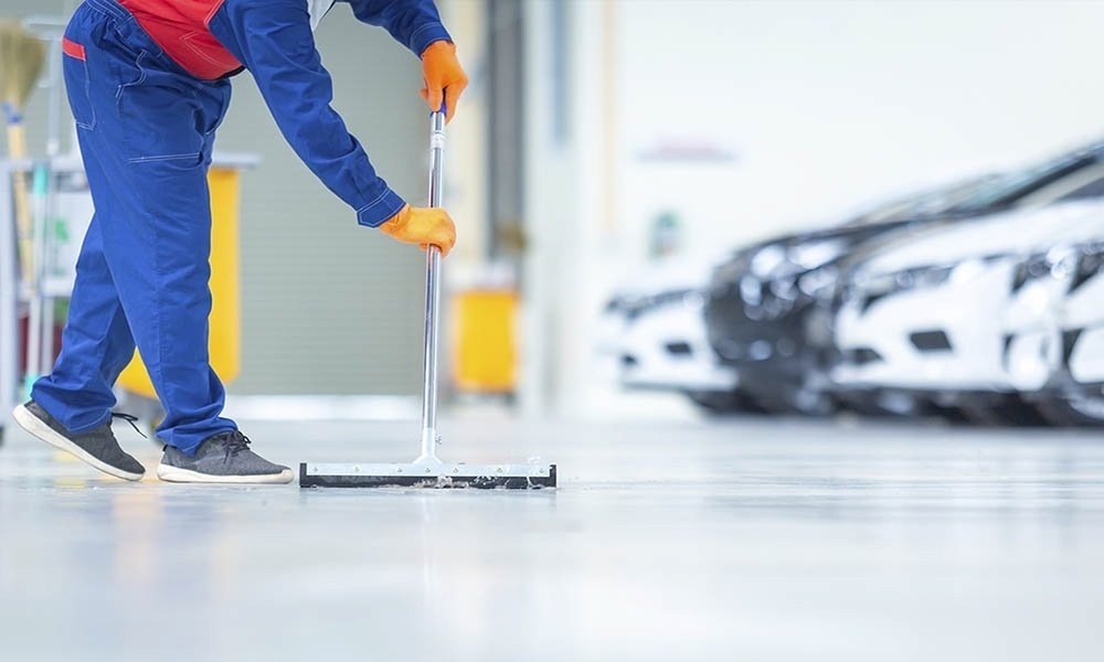 A man wearing orange gloves is performing car dealership cleaning by cleaning the floor of a car dealership.