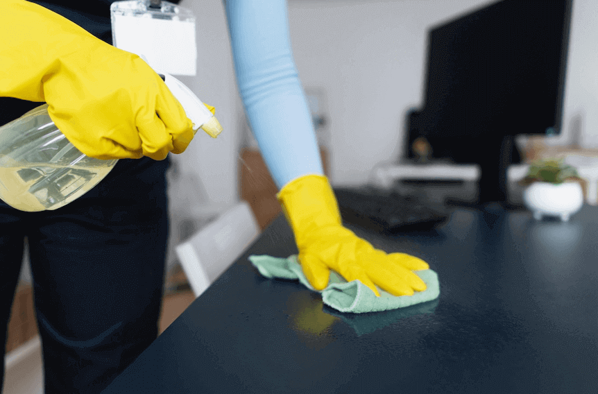 A person wearing yellow gloves cleans a desk as part of a commercial cleaning service.