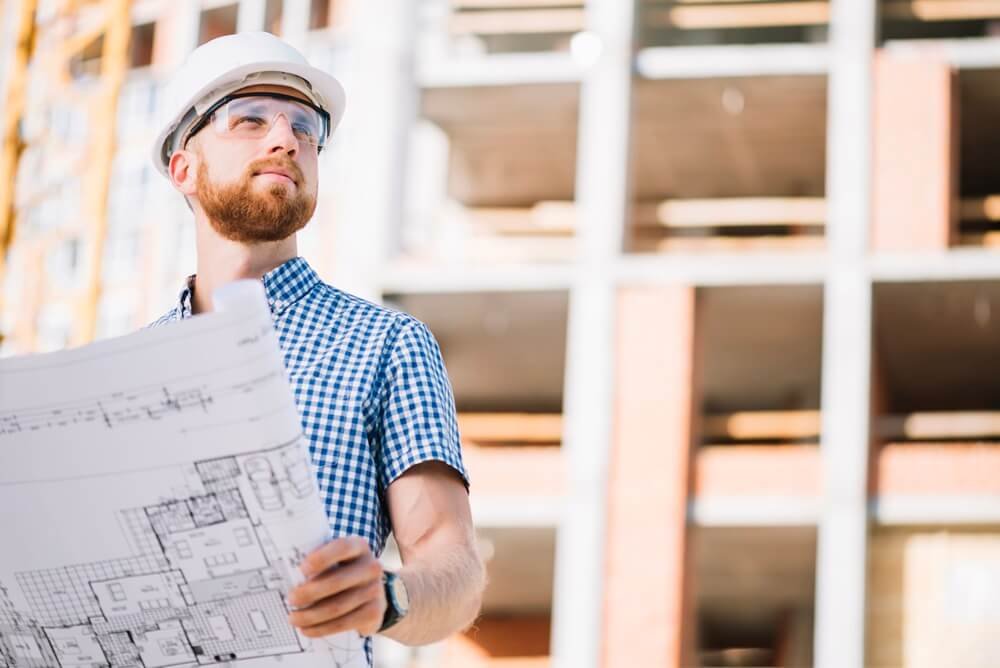 A man in a hard hat and glasses examines a blueprint, representing his work in general contracting.