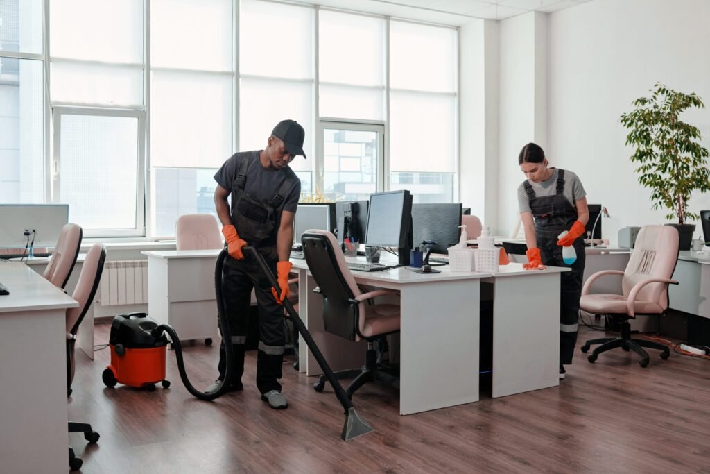 Two people offering office cleaning service by cleaning an office filled with computers and desks, ensuring a tidy and organized workspace.