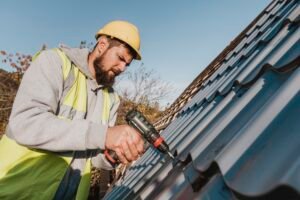 A bearded man in a hard hat uses a drill for roof repair, focused on his task in a construction setting.