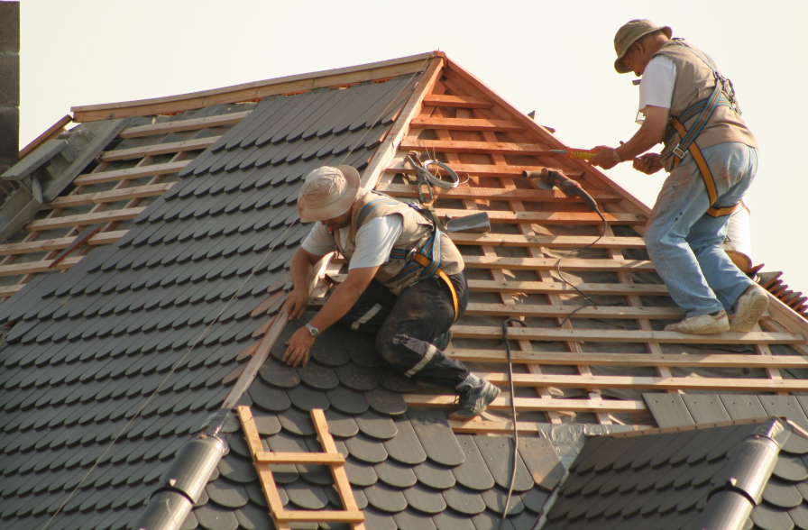 Two men repairing a roof, one on a ladder, focused on their work amidst a clear blue sky.