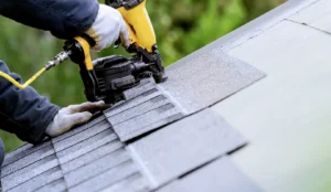 Our Blogs 11 Close-up of a roofer's hands in white work gloves using a yellow pneumatic nail gun to secure grey asphalt shingles onto a roof.