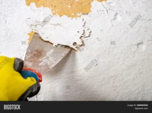 100local near 34 Professional paint removal process using a metal putty knife scraper to peel old, cracking white paint layers off a wall surface.