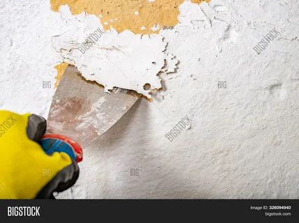 Professional paint removal process using a metal putty knife scraper to peel old, cracking white paint layers off a wall surface.