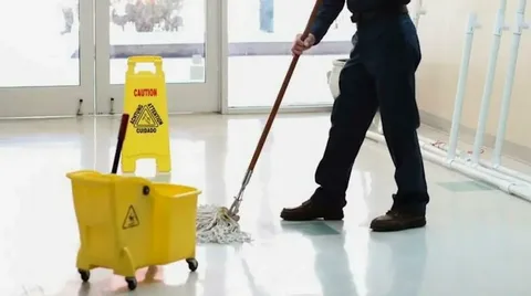 A maintenance worker mopping a bright commercial lobby floor next to a yellow caution sign, demonstrating professional porter services.